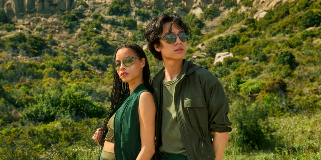 Two people wearing green-tinted lens Stoggles posed against a green forested hillside in casual outdoor clothing.