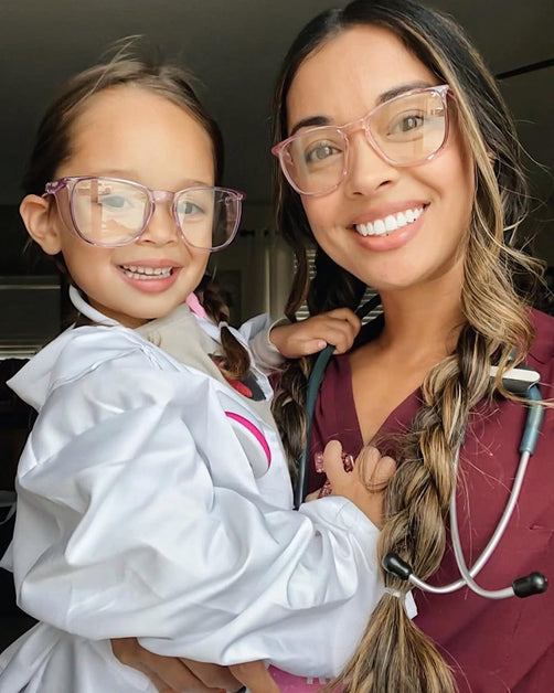 Woman holding a baby, both wearing Stoggles protective glasses, smiling together indoors.