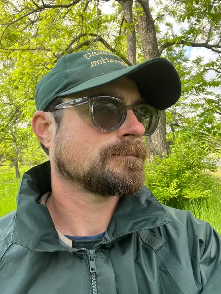 Man outdoors wearing tinted Stoggles safety glasses, a green cap, and a dark jacket.