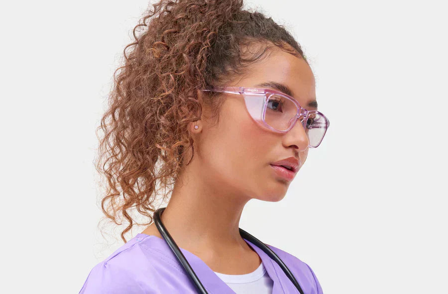 Model wearing translucent purple Stoggles safety glasses with side shields and purple scrubs, demonstrating wraparound eye protection and modern safety eyewear style.